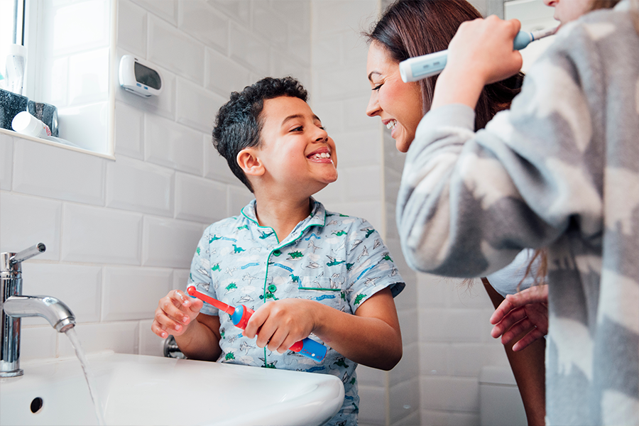 A young boy shows a woman his teeth by grinning widely. He is standing next to a bathroom sink and holding a toothbrush.