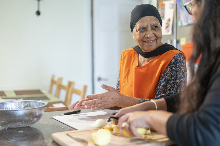 An older woman in an orange apron prepares food in a kitchen.