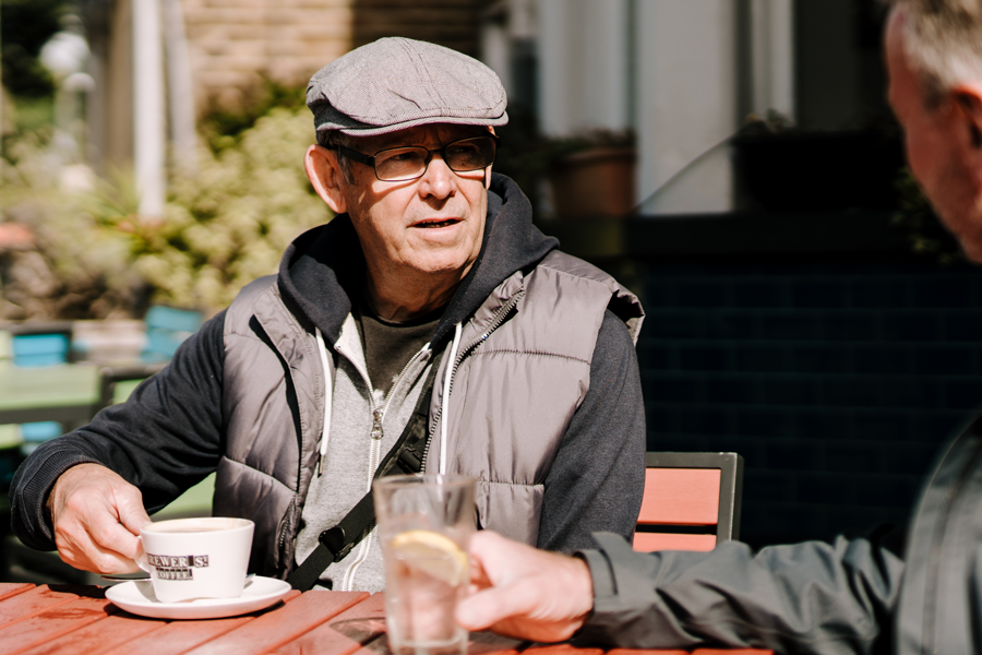 An older man wearing a flat cap and a gilet sits at an outdoor café table. He is talking to a companion and holding a coffee cup.