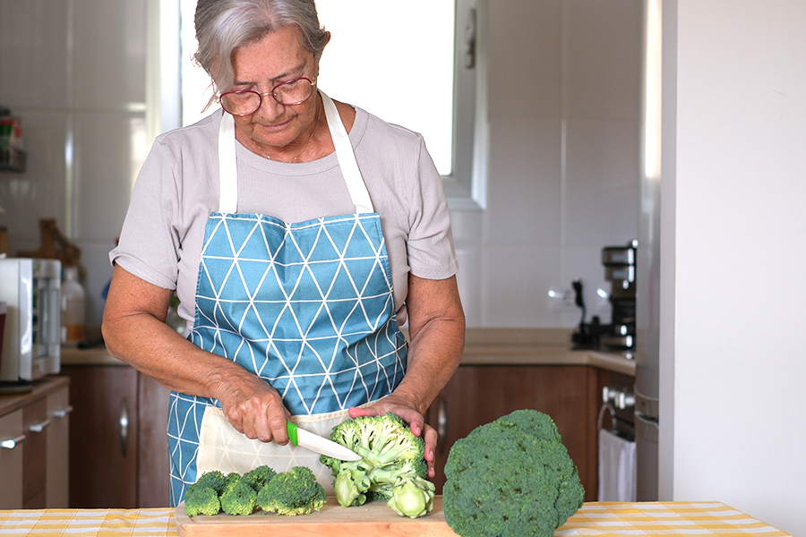 An older woman chops broccoli in her kitchen.