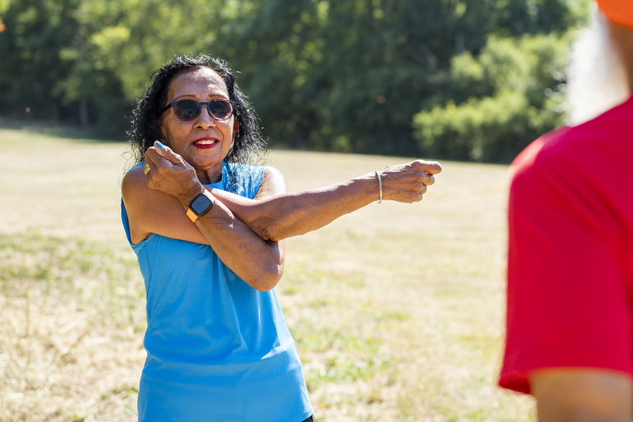 An older lady in a bright blue sports top stretching her arms in a sunny field.